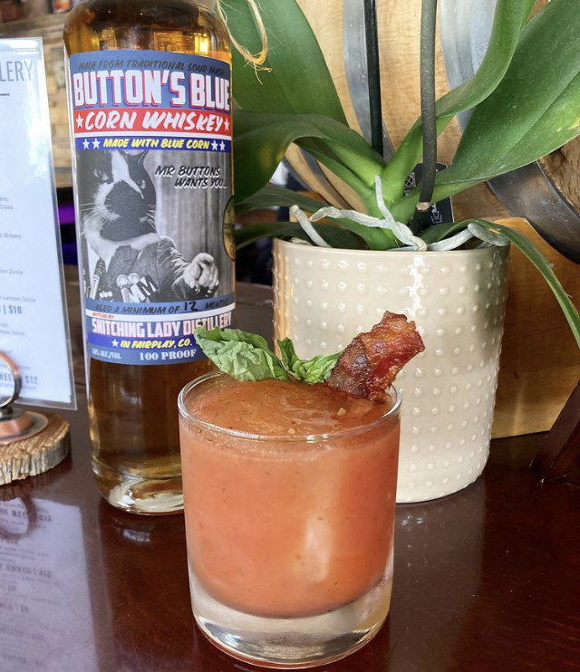 A bloody-style whiskey cocktail with basil and candied bacon beside the Button's Blue Corn bottle
