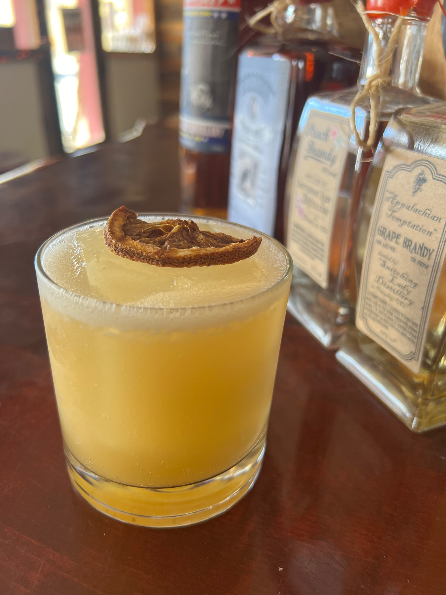 A citrus sour topped with a dried orange wheel beside Snitching Lady bottles