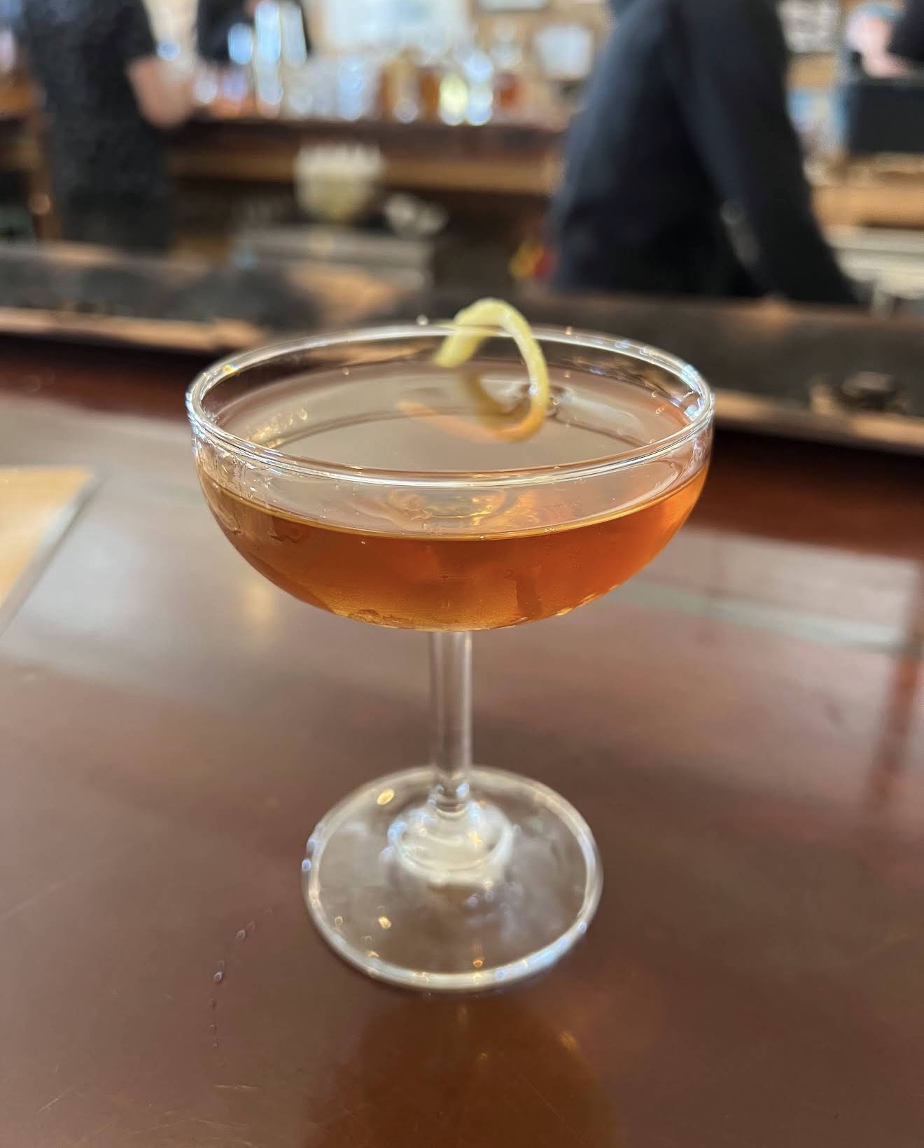 An amber Manhattan in a coupe with a lemon twist on the bar