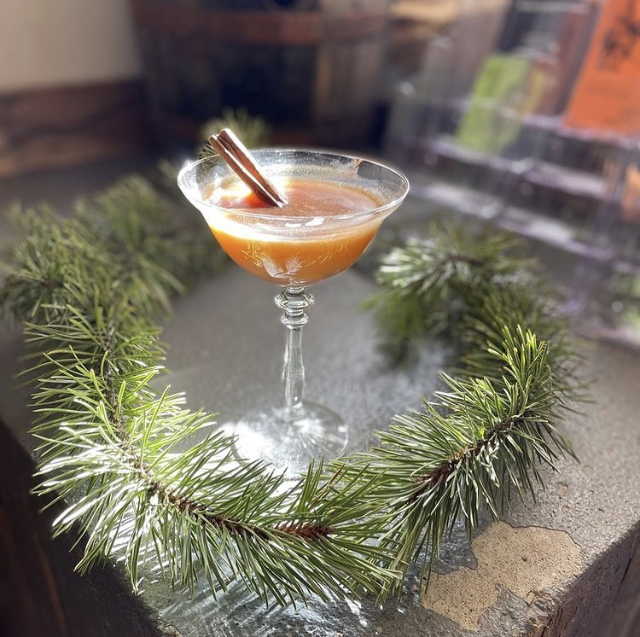 A spiced cider coupe with a cinnamon stick nested in a pine wreath