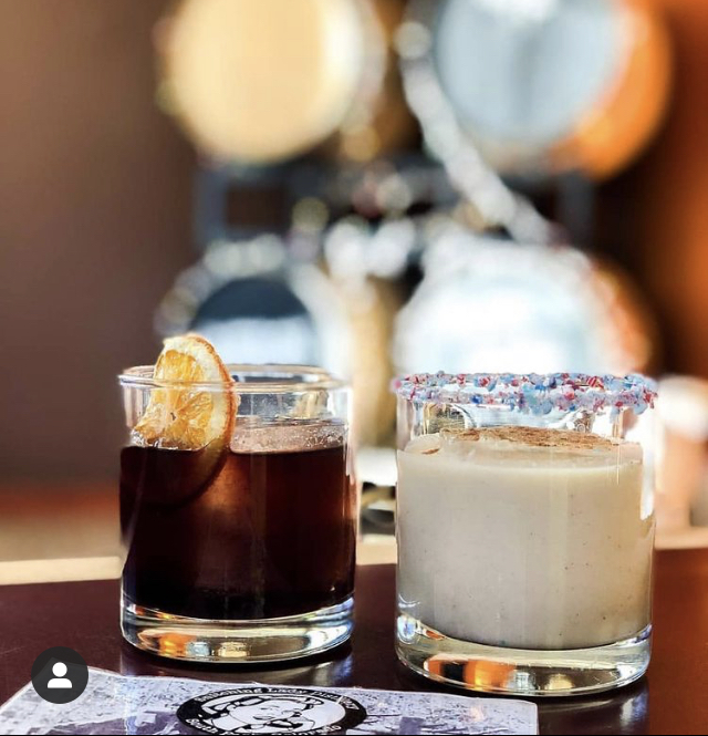 A dark citrus cocktail beside a creamy sprinkle-rimmed cocktail in front of the stills