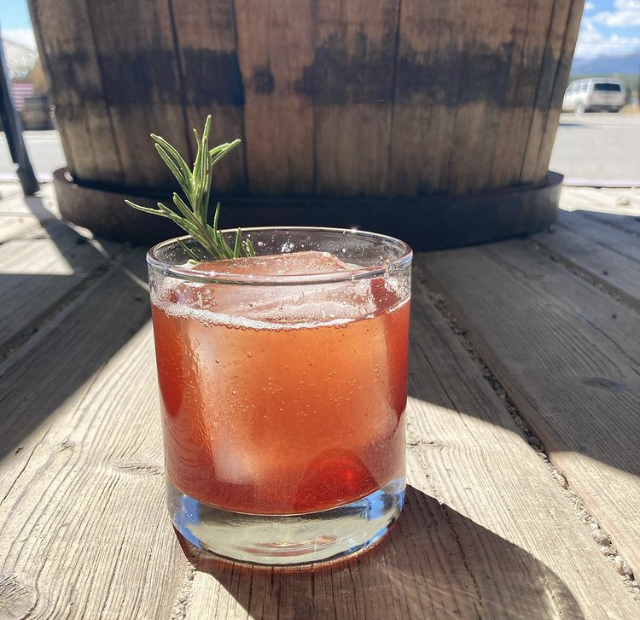 A ruby cocktail with a rosemary sprig on a sunny barrel-top table