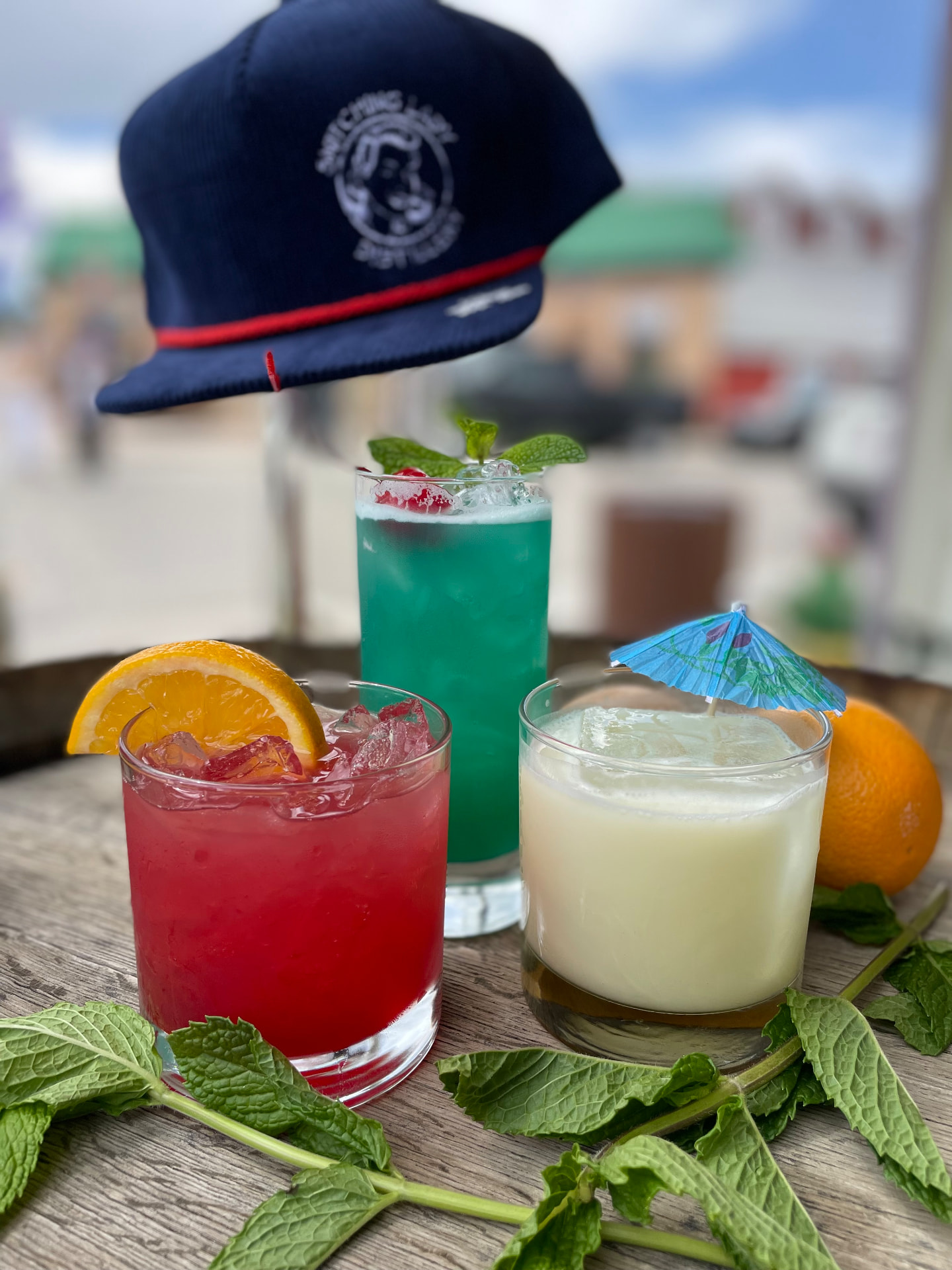 Three colorful cocktails with mint and citrus beside a Snitching Lady cap