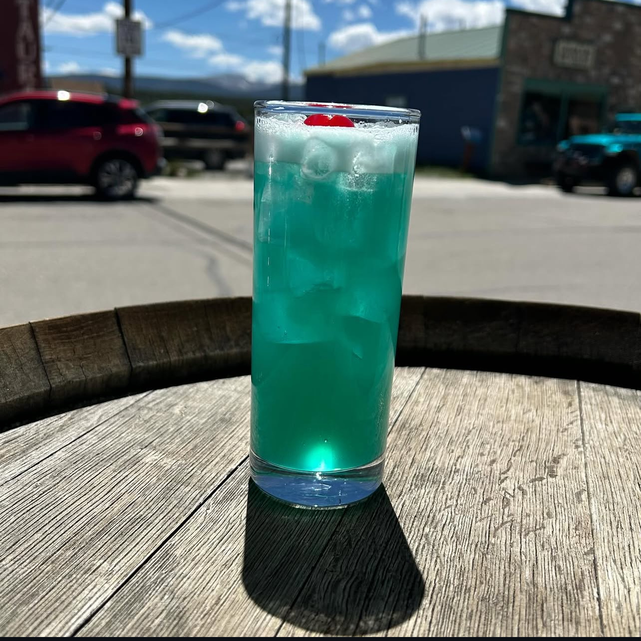 A bright turquoise highball with a cherry on a whiskey barrel outside the distillery