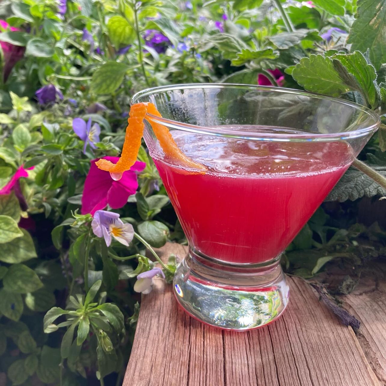 A bright pink cosmopolitan-style martini with an orange twist among garden flowers
