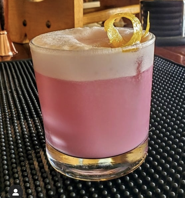 A pink sour with a thick foam cap and a curled lemon twist