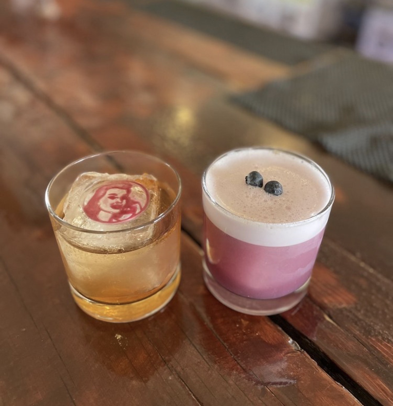 A logo-stamped old fashioned beside a blueberry sour on the reclaimed wood bar