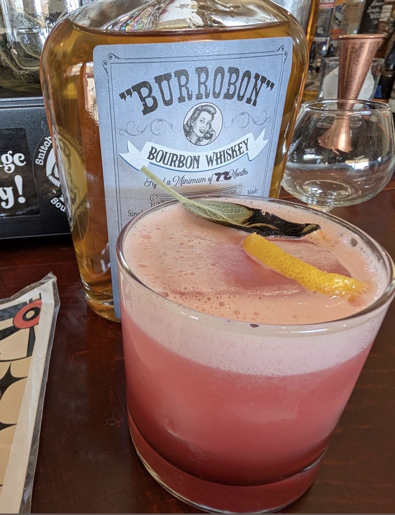 A pink sour with charred sage and lemon peel beside the Burrobon Bourbon bottle