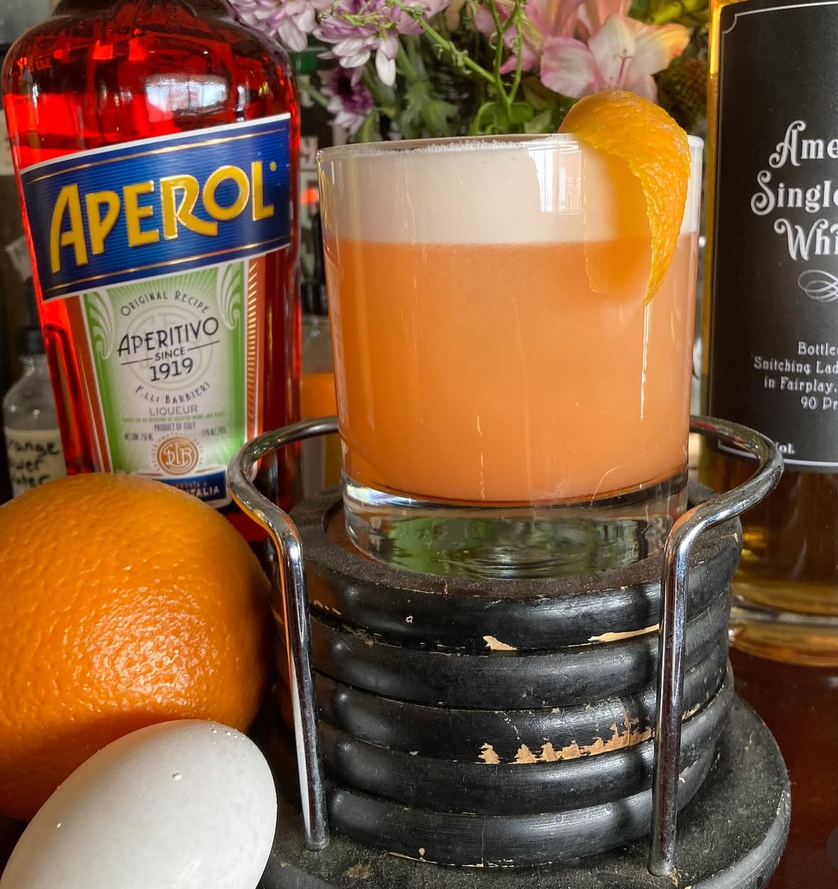 An Aperol whiskey sour with orange peel between the Aperol and American Single Malt bottles