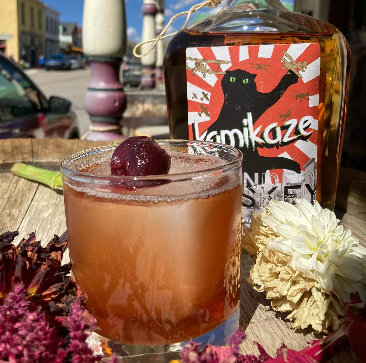 A cherry-topped Kamikaze Blend cocktail on the front porch with the branded bottle behind