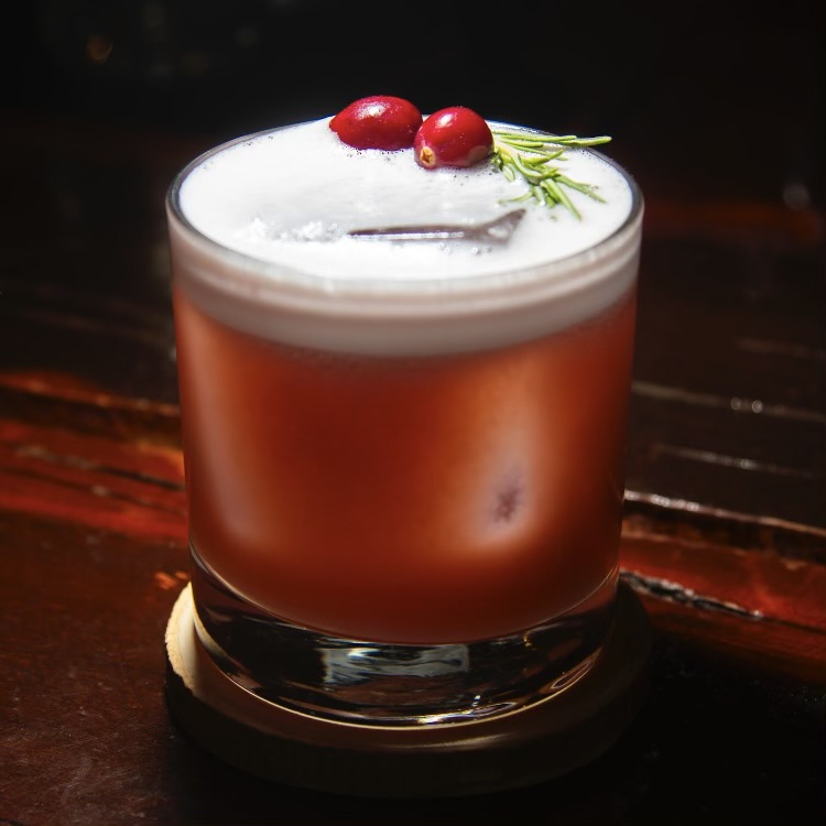 A cranberry whiskey sour with foam, fresh cranberries and rosemary