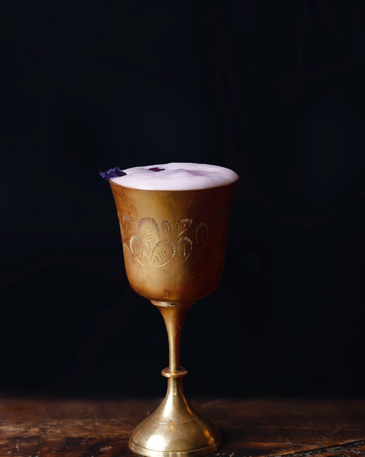 A violet gin fizz in an etched brass goblet against a dark backdrop