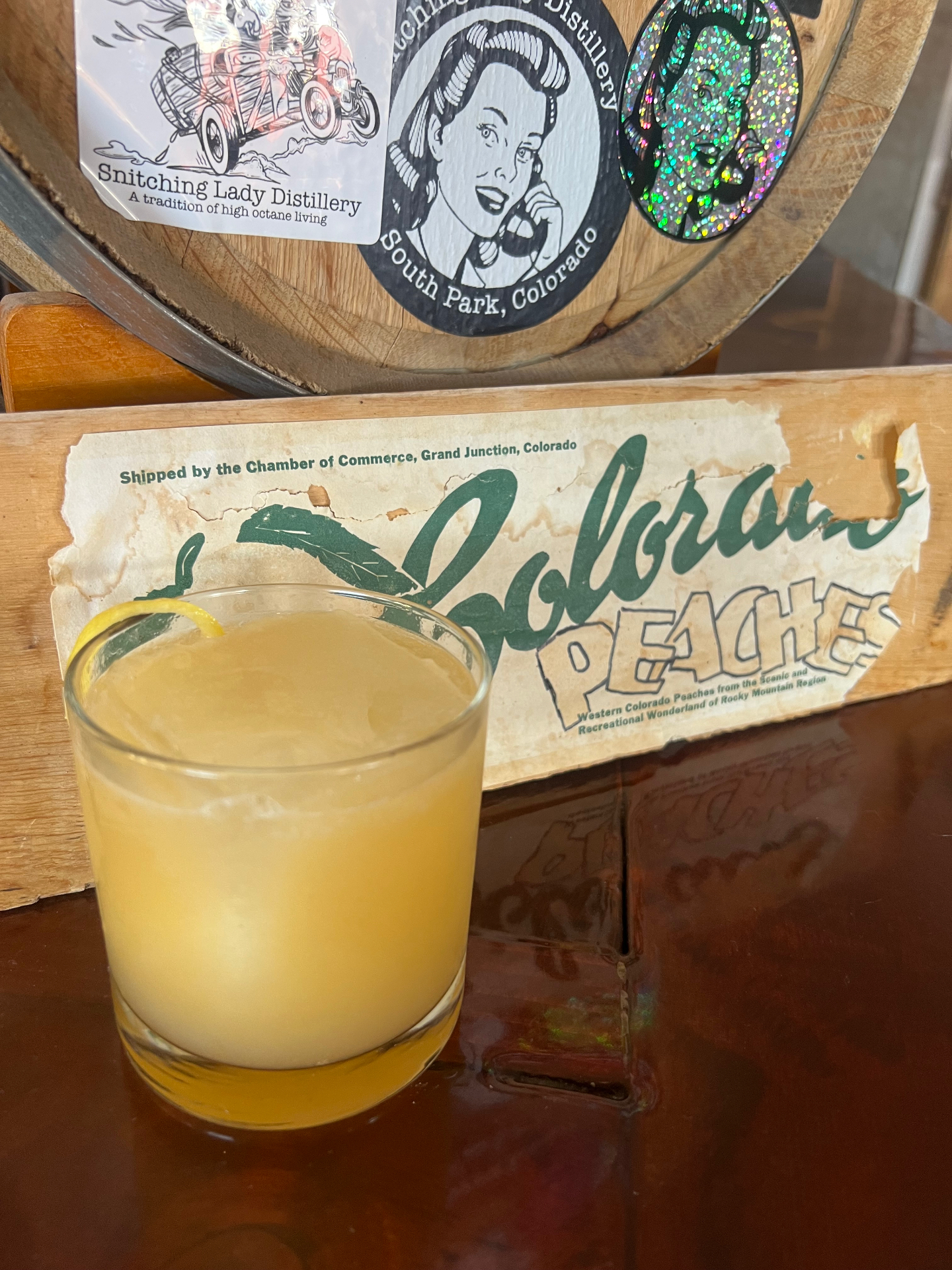 A Colorado peach whiskey sour with a lemon twist beside a vintage Colorado Peaches crate
