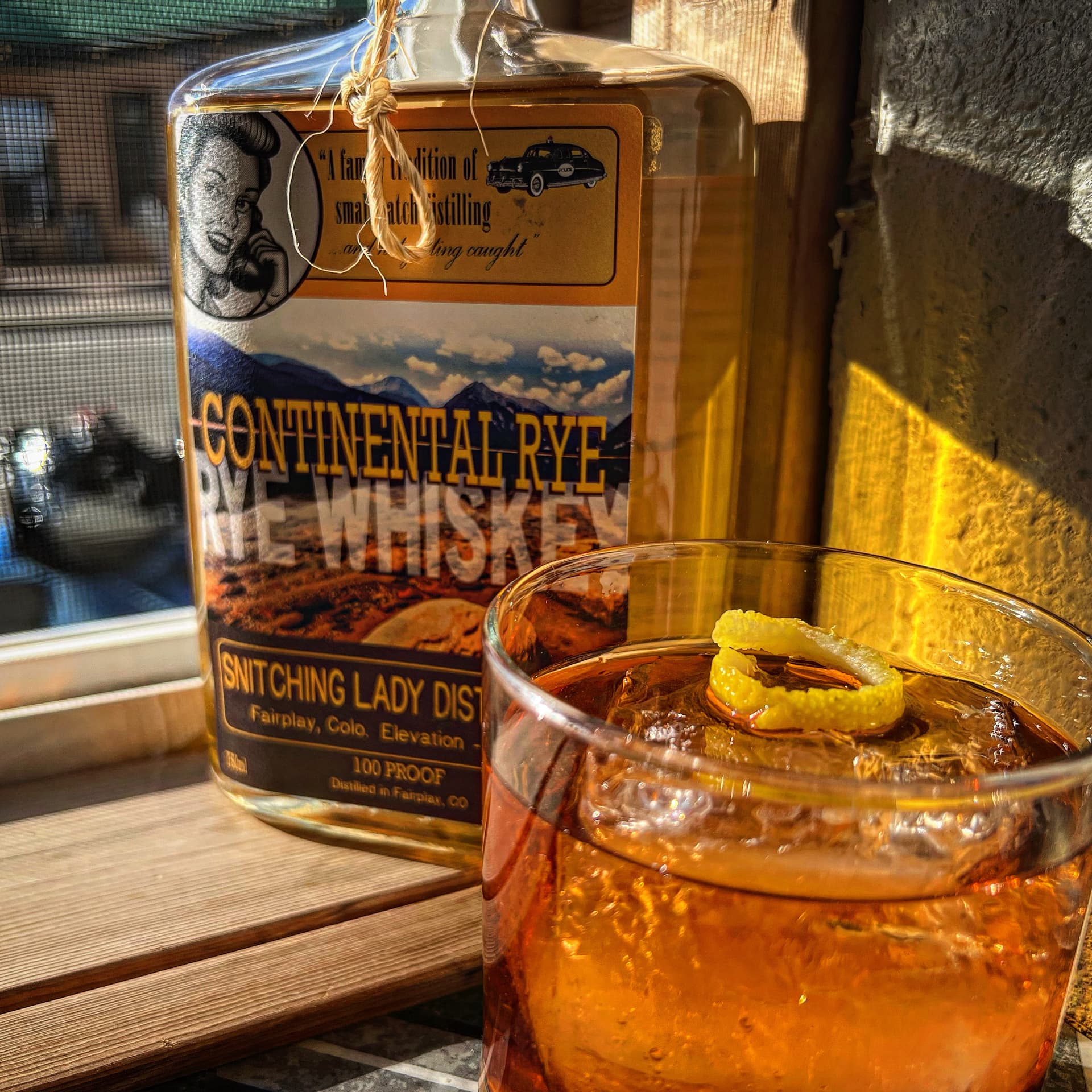 An old fashioned with a lemon twist beside a Continental Rye bottle in golden light