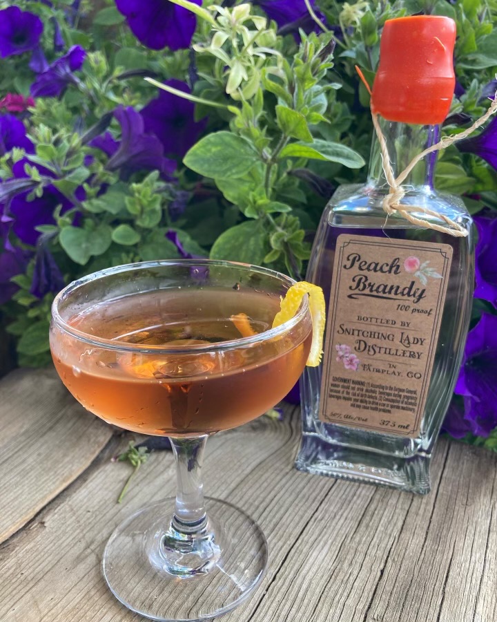 A peach brandy Manhattan in a coupe with a lemon twist beside the Peach Brandy bottle