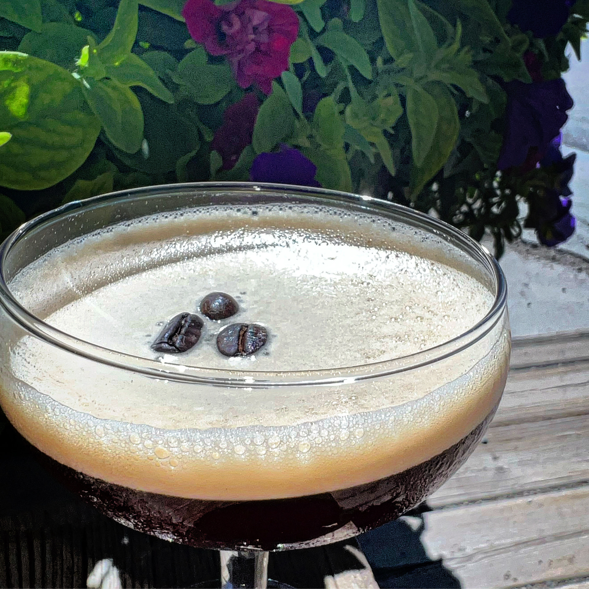 An espresso martini topped with three coffee beans among garden flowers