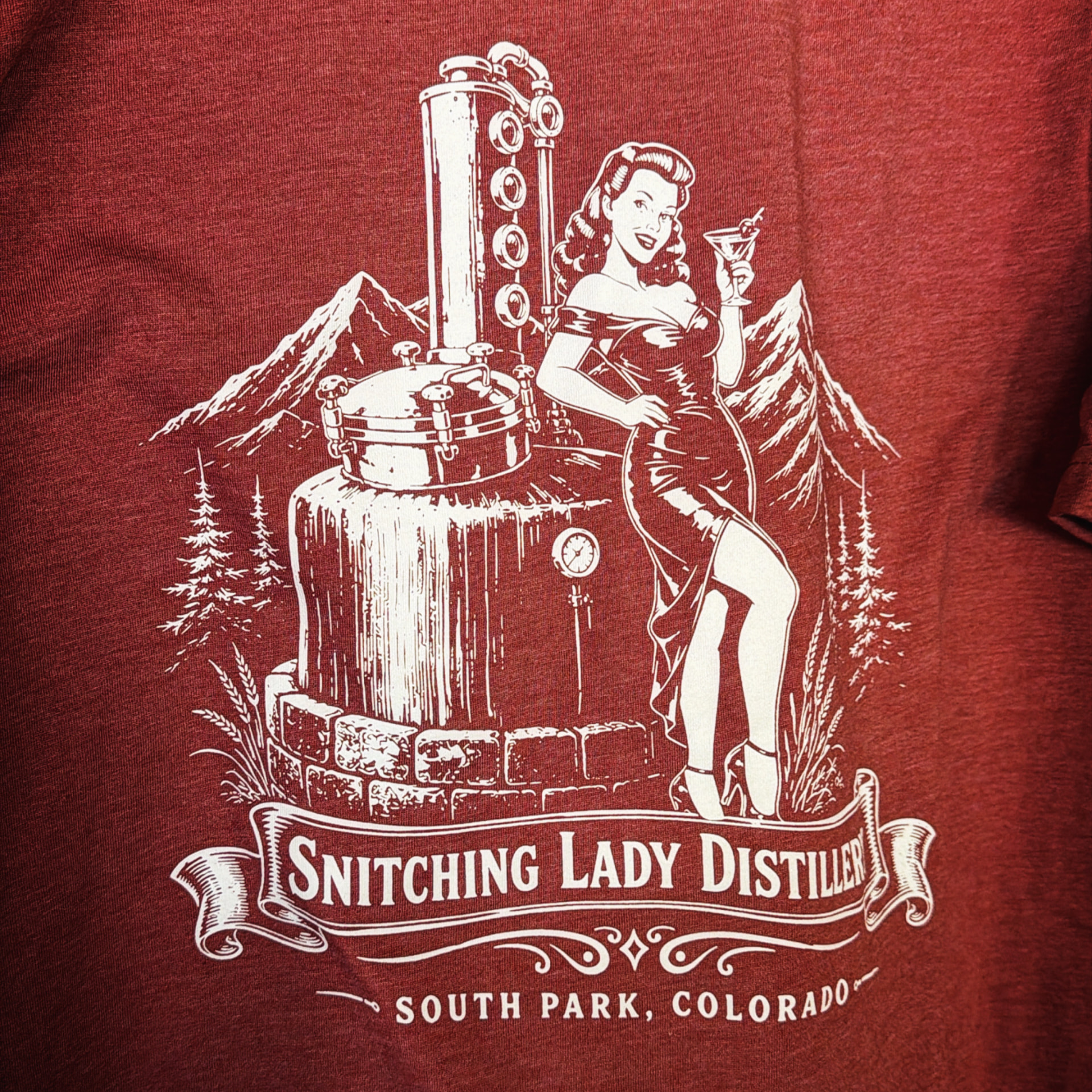 Maroon tee with the vintage Snitching Lady Distillery mountain still logo in white