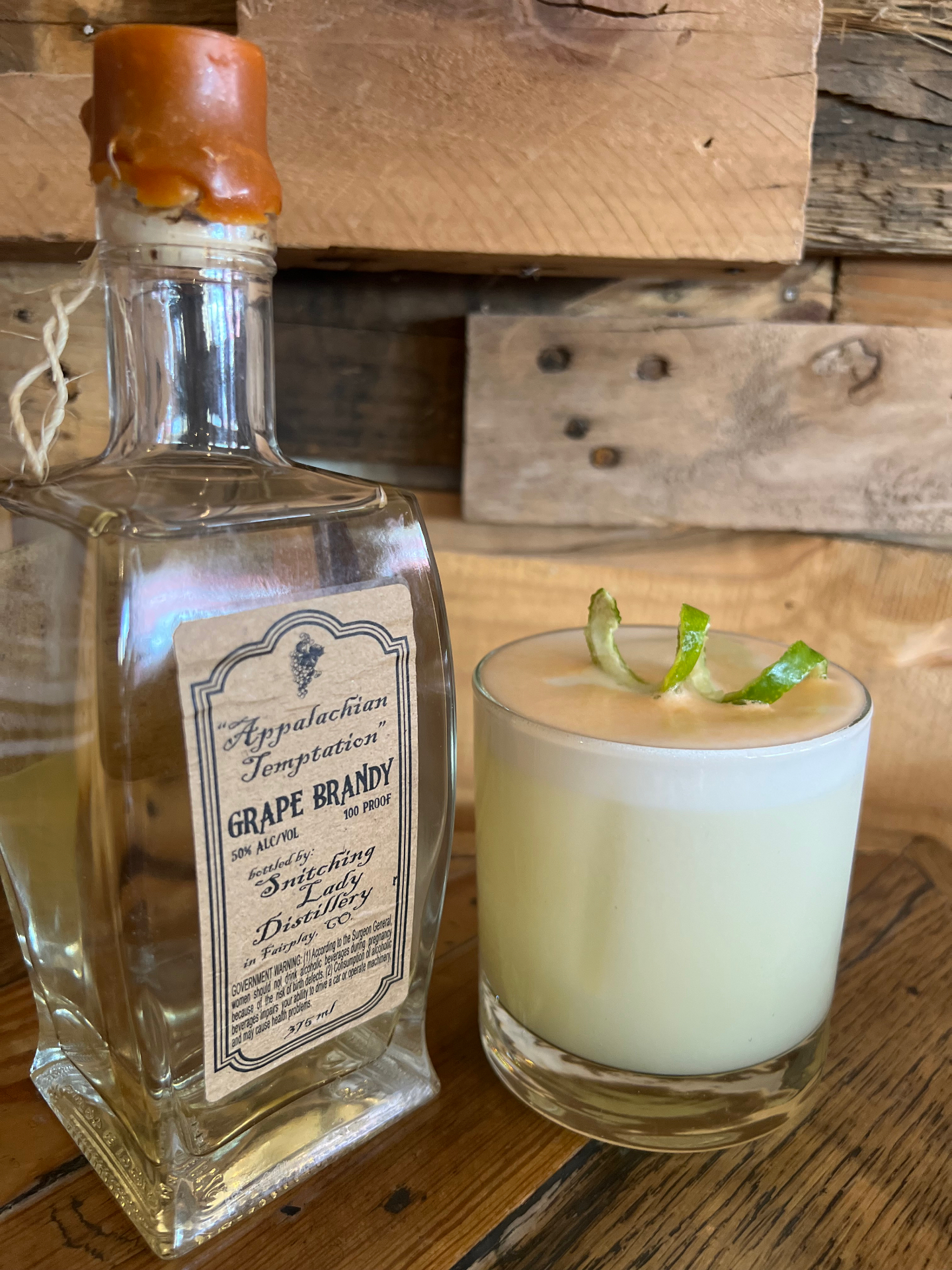 A frothy grape brandy sour beside the Appalachian Temptation Grape Brandy bottle