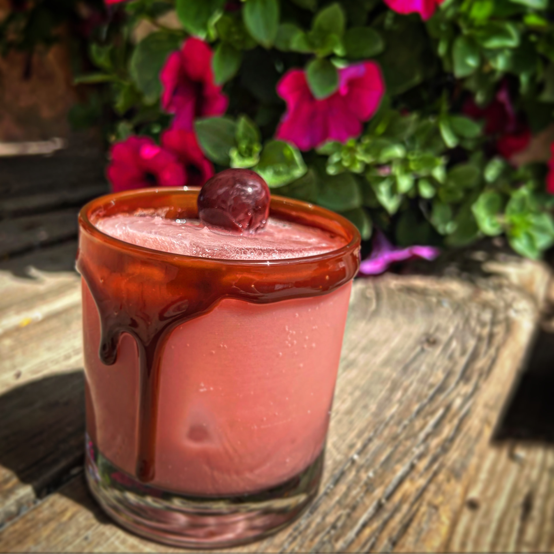 A cherry cream cocktail with a syrup-drizzled rim beside bright petunias