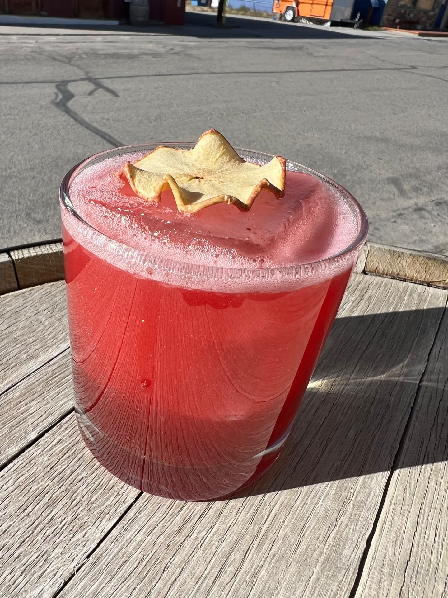 A crimson cocktail with a dried apple garnish on a sunny barrel-top table
