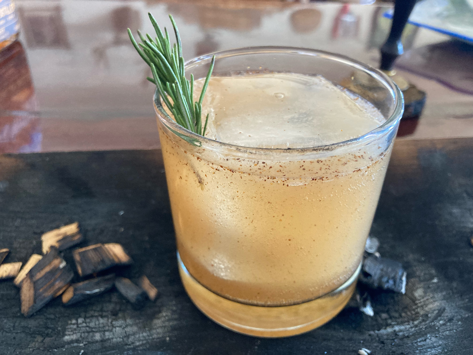 A smoked whiskey cocktail over a large ice cube with a rosemary sprig and charred oak chips