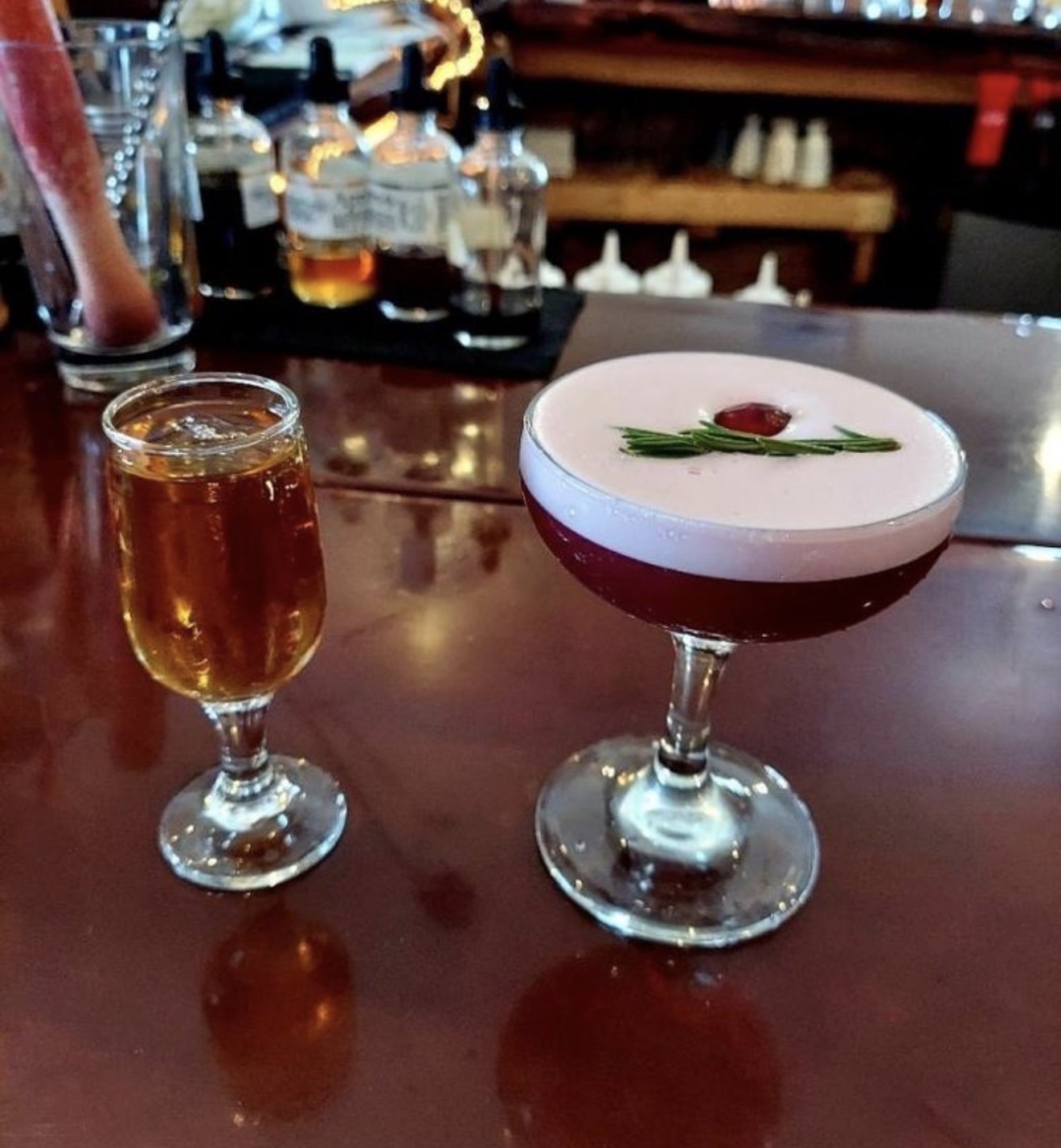 A cranberry rosemary sour beside a whiskey pour on the bar