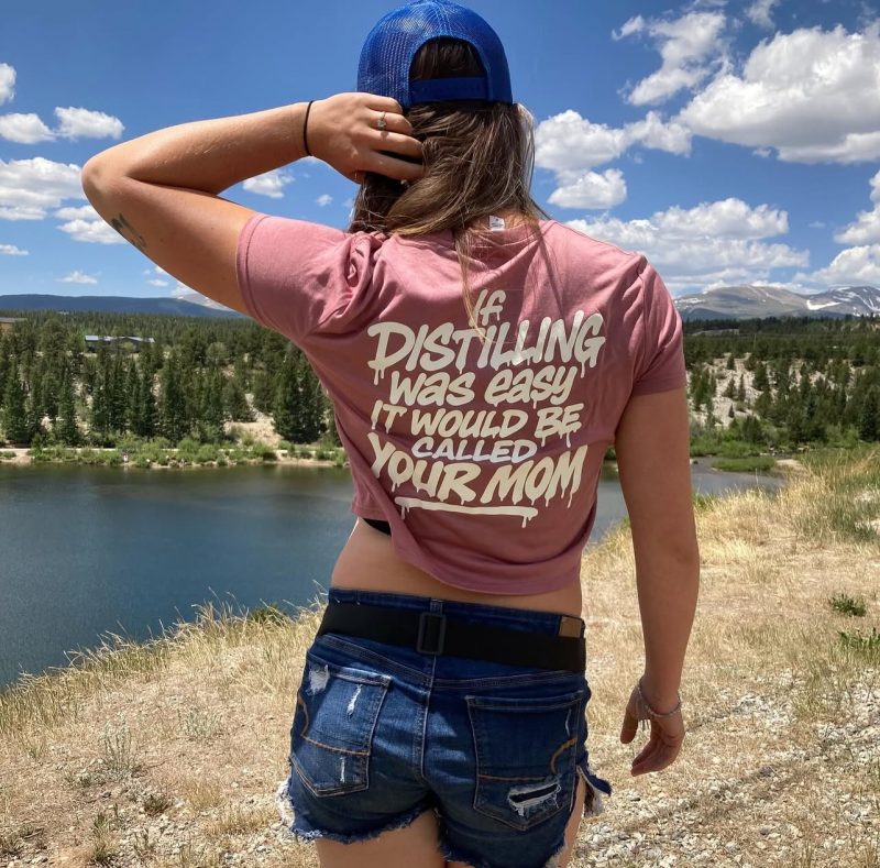 Rose t-shirt with the back print 'If distilling was easy it would be called your mom', worn by a mountain lake