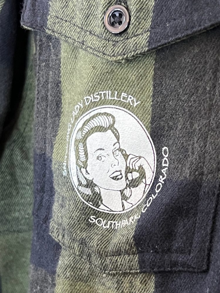 Olive-plaid flannel with the round Snitching Lady Distillery South Park, Colorado crest on the chest