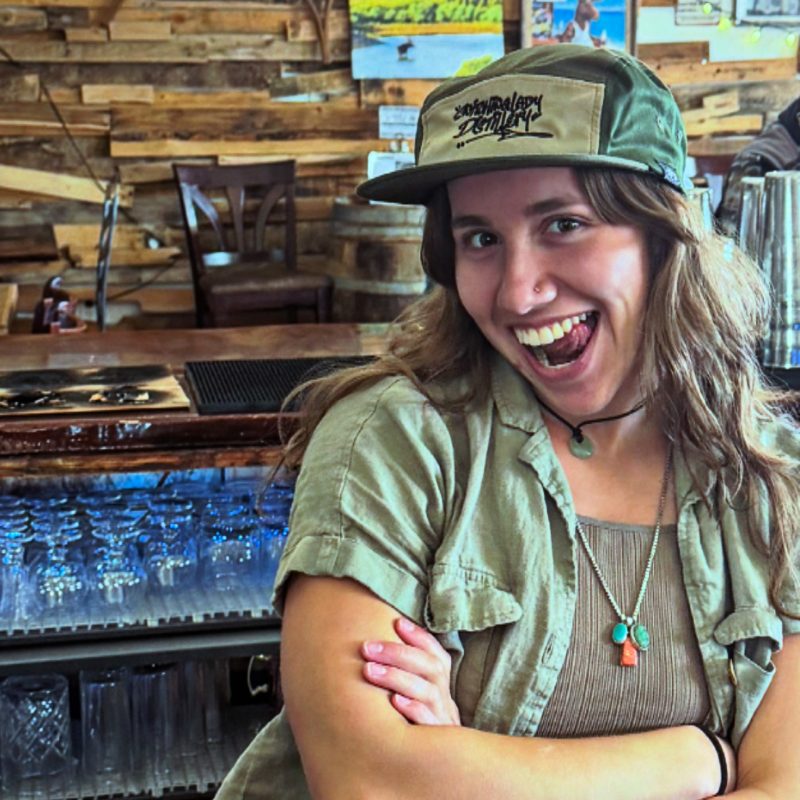 A bartender wearing an embroidered Snitching Lady Distillery snapback cap behind the bar