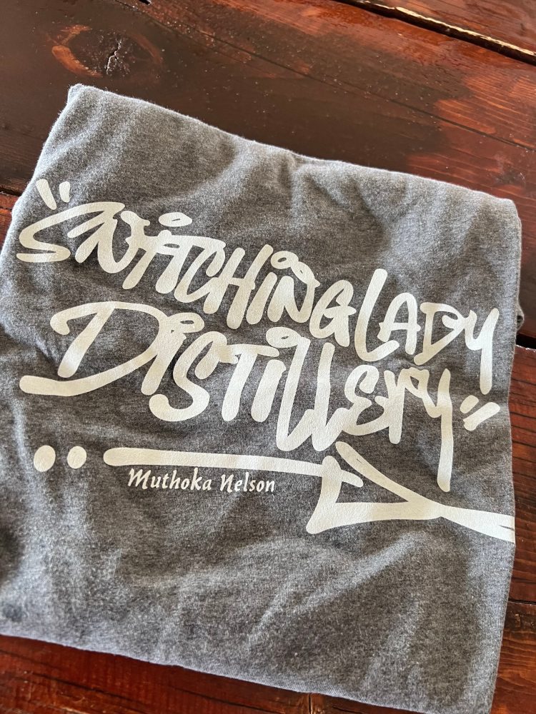 Heather-gray tee with the Snitching Lady Distillery graffiti-script logo, signed by artist Muthoka Nelson
